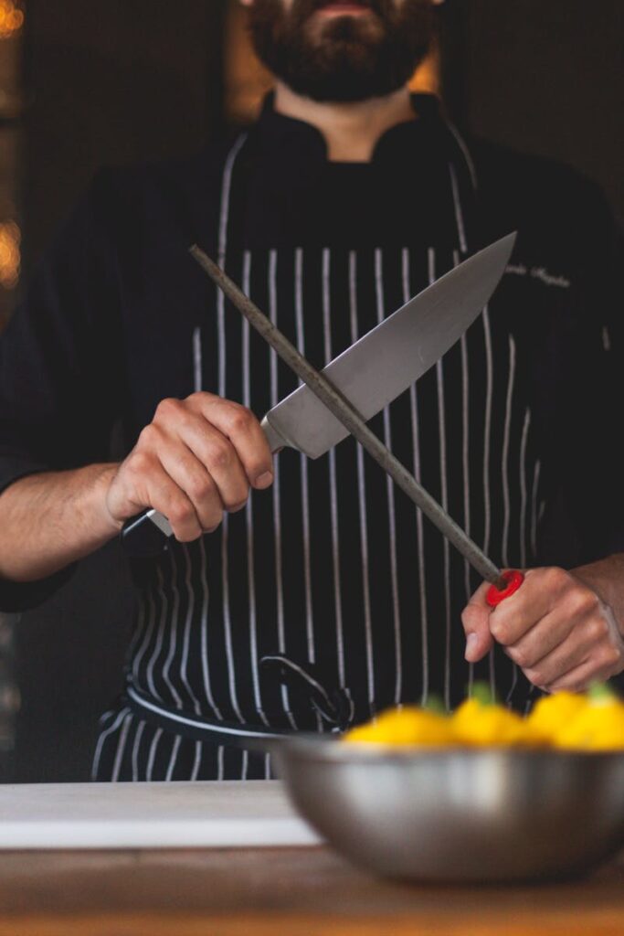 A chef in striped apron sharpening a knife with a honing steel in a professional kitchen.